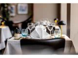 Et pænt dækket restaurantbord med foldede hvide servietter, vinglas, farverige vandglas, bestik og en lille sort lampe. Baggrunden er blødt sløret.