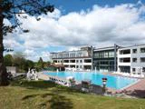 Et moderne hotel med store vinduer har udsigt til en rektangulær udendørs pool omgivet af liggestole, grønt græs, træer og en delvis overskyet himmel.