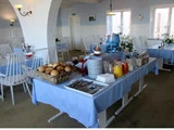 Morgenmadsbuffet med rundstykker, drikkevarer, tallerkener og bestik på en blå dug i en lys spisestue med hvide stole og indretning.