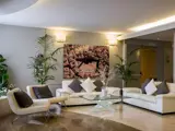 Moderne stue med hvide sofaer, sofabord i glas, grønne planter og et stort luftfoto af byen på væggen. Neutrale toner og blød belysning skaber en hyggelig, elegant atmosfære.