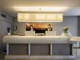 To receptionister står bag en moderne hotelreception og arbejder ved computere. En stor lampe hænger ovenover, og blomster pynter på disken.