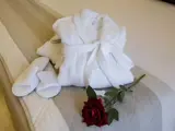 En sammenfoldet hvid badekåbe, hvide tøfler og en rød rose er sirligt arrangeret på en beige sengeløber på en opredt seng.