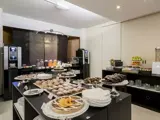 Morgenmadsbuffet med bagværk, kager, frugt, juicedispensere, kaffemaskiner og stablede tallerkener i et lyst, moderne spiseområde.