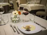 Et restaurantbord dækket med en desserttallerken, et glas rødvin, vandglas, sølvtøj og en vase med hvide og orange blomster.
