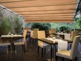 Udendørs restaurantterrasse med beige stole og borde til spisning, hvide duge, vinglas, stearinlys og en beige udtrækkelig markise; grønne planter i baggrunden.