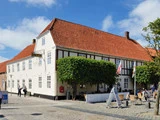 En historisk hvid bygning med rødt tegltag, udeservering og grønne træer på en brostensbelagt gade under en blå himmel.