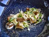 En gourmetret med tynde spåner af ost, friske krydderurter, sprødt brød og skiveskårne grøntsager kunstfærdigt anrettet på en mørkeblå keramiktallerken.