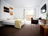 Et moderne hotelværelse med dobbeltseng, skrivebord, stol, væghængt tv og åbne døre, der fører ud til en gårdhave med træer, der kan ses udenfor. Værelset har minimal indretning og neutrale farver.