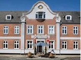 Et hotel i røde mursten med hvide kanter, høje vinduer, en dekorativ gavl og udendørs siddepladser under en blå parasol med to danske flag ved indgangen.