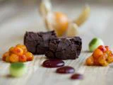 To stykker chokoladebrownie er omgivet af orange bær, grønne dråber og rød sauce på en træplade.