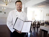 En kok i hvid uniform står i en restaurantspisestue, smiler og holder en åben menu op mod kameraet.