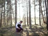 En person iført et sort forklæde knæler på skovbunden og undersøger jorden, omgivet af høje træer og sollys, der filtrerer gennem grenene.