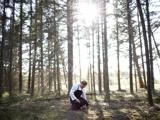 En person i hvid skjorte og sort forklæde knæler på skovbunden, omgivet af høje træer med sollys, der strømmer gennem grenene.