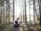 En person iført et sort forklæde knæler på skovbunden og undersøger jorden, omgivet af høje træer og sollys, der filtrerer gennem grenene.