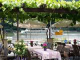 Udendørs café med borde og stole under en grøn pergola, potteblomster på bordene og en flod med en forbipasserende båd i baggrunden.