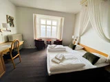 Et lyst hotelværelse med en dobbeltseng, to grønne puder, et skrivebord med en stol, to lænestole ved et stort vindue og et lille rundt bord.