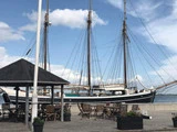 Et højt skib med tre master ligger til kaj ved havnefronten ved siden af en gårdhave med tomme træborde og stole under et lysthus.