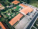 Luftfoto af bygninger med orange tage, et anlagt grønt område, parkeringsplads med biler og tennisbaner i baggrunden.