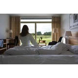 En kvinde i hvid kåbe sidder på en seng foran et stort vindue og kigger ud på et grønt landskab med træer og et bord, der er dækket til morgenmad.