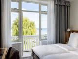 Et lyst hotelværelse med en pænt redt seng vender ud mod store vinduer og en balkon med udsigt over træer og en fjern udsigt over havet.