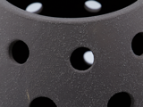 Nærbillede af et struktureret, mørkegråt objekt med runde huller og en ru, plettet overflade. Hvidt lys er synligt gennem hullerne i baggrunden.