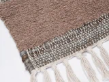 Et nærbillede af et vævet brunt og beige tekstil med frynsede kvaster langs kanten, vist på en hvid baggrund.