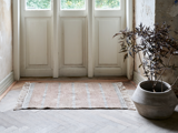 Et lille beige tæppe med frynsede kanter ligger foran hvide dobbeltdøre ved siden af en stor potteplante med mørke blade på et trægulv.