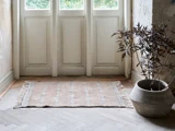 Et lille beige tæppe med frynsede kanter ligger foran hvide dobbeltdøre ved siden af en stor potteplante med mørke blade på et trægulv.