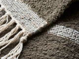 Nærbillede af et vævet stof med tykke, strukturerede fibre i jordfarver og med knyttede frynser på den ene kant.