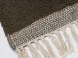 Nærbillede af et vævet tekstil med en mørkebrun struktureret del, et lysere vævet område og cremefarvede frynsede kvaster langs kanten.