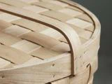 Nærbillede af en flettet trækurv med en buet træhank og synlige metalklammer, der fastgør strimlerne.