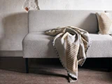 En lysegrå struktureret sofa med et mønstret beige og brunt tæppe over sig og en matchende pude mod en slidt, falmet væg.