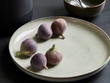 Fem friske figner med lilla-grønt skind er arrangeret på en stor, rund, råhvid keramiktallerken, der er placeret på en mørk overflade.