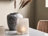 En stor grå keramikvase med tørret pampasgræs ved siden af en lysestage i matteret glas på et marmorbord med en kurvestol i baggrunden.