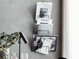 En væghængt magasinholder med flere magasiner og tidsskrifter ved siden af et skrivebord med en sort lampe, en plante og et glas.