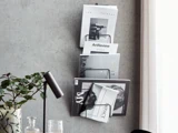 En væghængt magasinholder med flere magasiner og tidsskrifter ved siden af et skrivebord med en sort lampe, en plante og et glas.