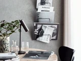 En moderne arbejdsplads med et træbord, en sort lampe, en plante i en vase, et glas vand og magasiner på en væghængt reol mod en grå væg.