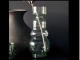 En klar, struktureret glasvase med smal hals og afrundet krop står på en mørk overflade mod en mørk baggrund. Et tørret blad er placeret inde i vasen.
