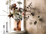 En glasvase med tørrede efterårsblomster står i vindueskarmen ved siden af to lysestager, den ene med et hvidt stearinlys, mod en struktureret beige væg.