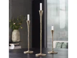 Tre metalliske lysestager i forskellige højder med hvide stearinlys placeret på et mørkt bord. En vase med blomster og stablede bøger er i den slørede baggrund.