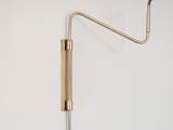 Minimalistisk væghængt lampearmatur i messing med en lang arm og to hængsler, fastgjort til en hvid væg.
