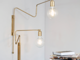 En moderne væglampe i messing med synlige pærer og justerbare arme er monteret på en hvid væg ved siden af en stak bøger og magasiner.