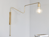En moderne vægmonteret lampe i guld med en synlig pære strækker sig ud over en minimal hvid væg, hvor en del af en bog er synlig i nederste højre hjørne.