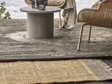 Et vævet tæppe ligger på en træterrasse med et rundt bord, kurvestole og et stribet tæppe, der er draperet over en stol nær et vandløb.