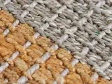 Nærbillede af et vævet tæppe med tykke, strukturerede fibre i to farver: naturbrun og lysegrå, arrangeret i et kurvefletningsmønster.
