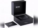 En sort Hugo Boss-urkasse med åbent låg, der viser et polstret rum indeni og et hæfte med påskriften BOSS placeret ved siden af.