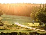 En person spiller golf på en frodig, solbeskinnet bane omgivet af træer med gyldent sollys, der strømmer gennem skoven i baggrunden.