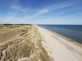 En sandstrand omgivet af græsklædte klitter strækker sig langs et roligt blåt hav under en klar, solrig himmel.
