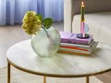 Et rundt sofabord i marmor med en tekstureret glasvase med en hortensia, en stak bøger og et tændt tofarvet stearinlys i en holder.