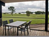 Udendørs terrasse med et metalbord og fire stole på et trædæk med udsigt over en græsmark, træer og en sø under en overskyet himmel.
